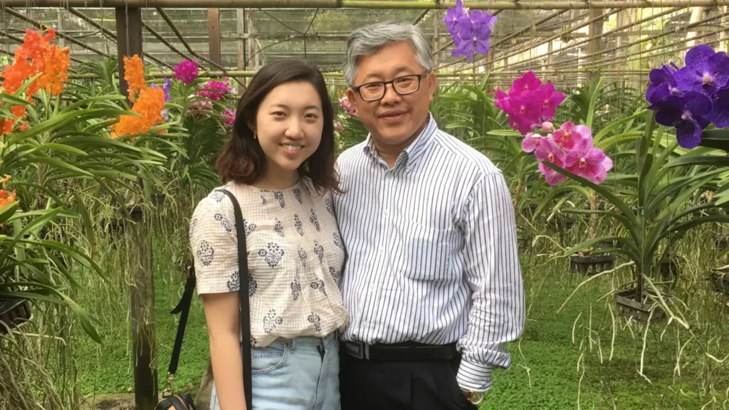 Two people, a younger daughter and an older father from China. They are standing together in front of some colourful flowers during the day