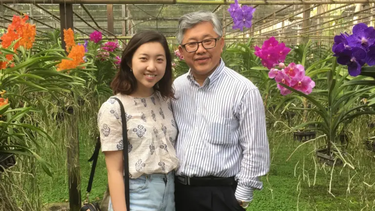 Two people, a younger daughter and an older father from China. They are standing together in front of some colourful flowers during the day